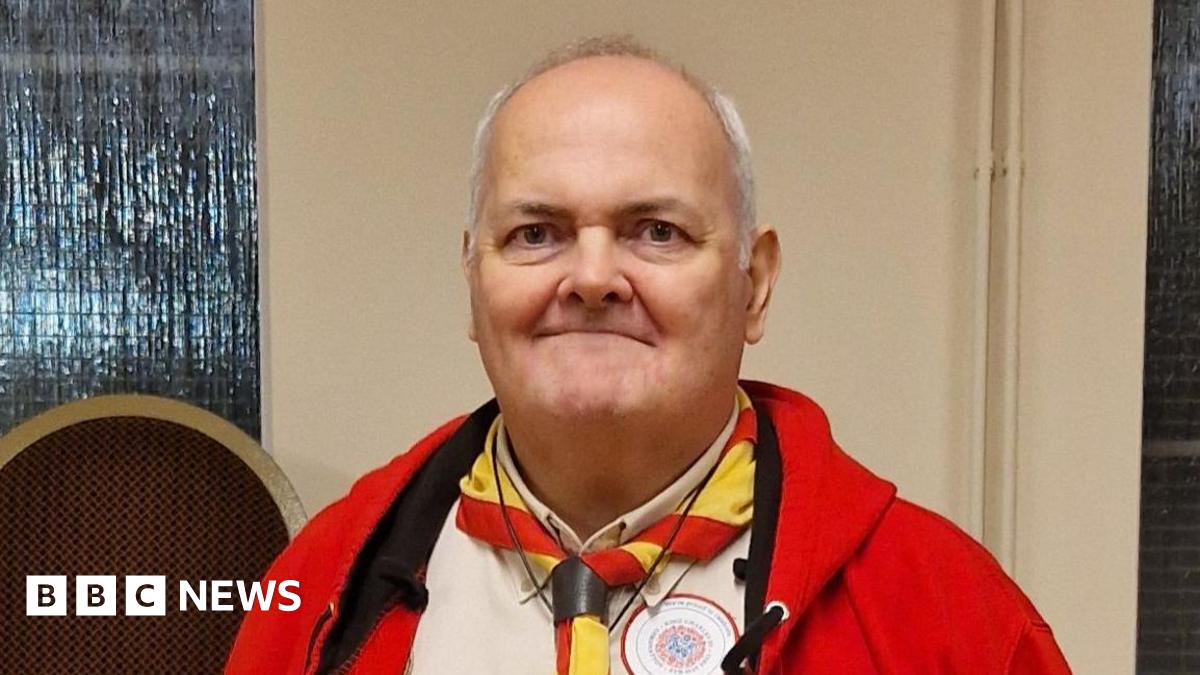 Matthew Padley with white hair wearing a Scout leader uniform complete with red fleece beige shirt and red and yellow necker. He is smiling.