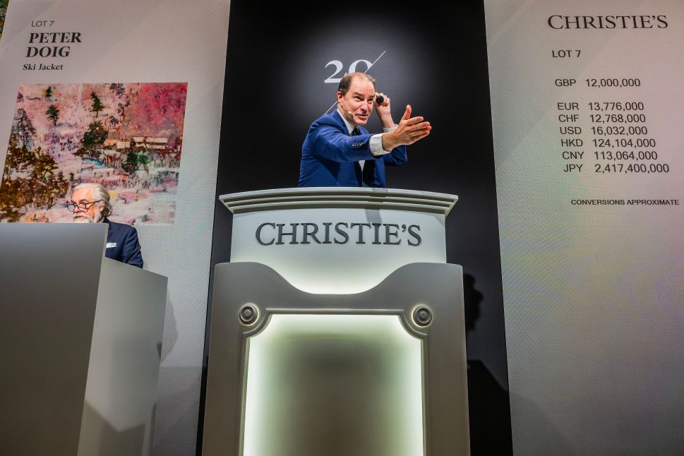 Auctioneer gestures from the Christie’s podium during the sale of Peter Doig’s Ski Jacket, with the painting and multi-currency price list displayed on large screens behind him.