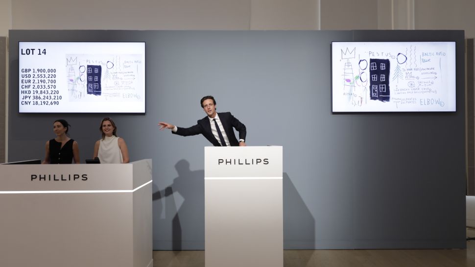 A Phillips auctioneer points to the room beside screens displaying Jean-Michel Basquiat’s Pestus and its current bids in multiple currencies.
