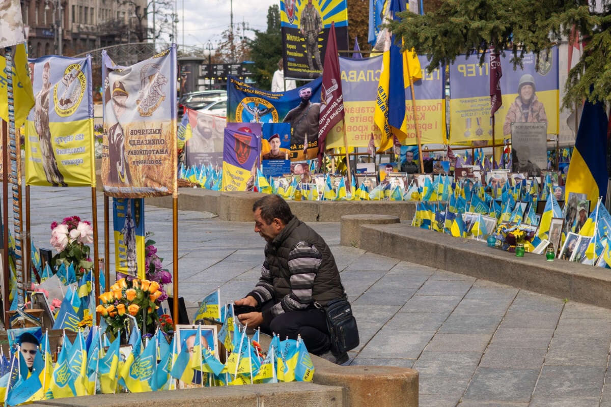 A man kneels in the middle of a memorial covered in Ukrainian flags.