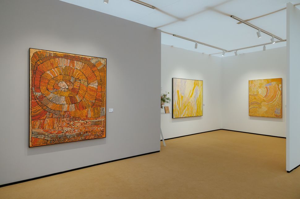 Gallery booth featuring large Aboriginal paintings in warm yellow and orange earth tones, each with intricate linear patterns hanging on neutral walls
