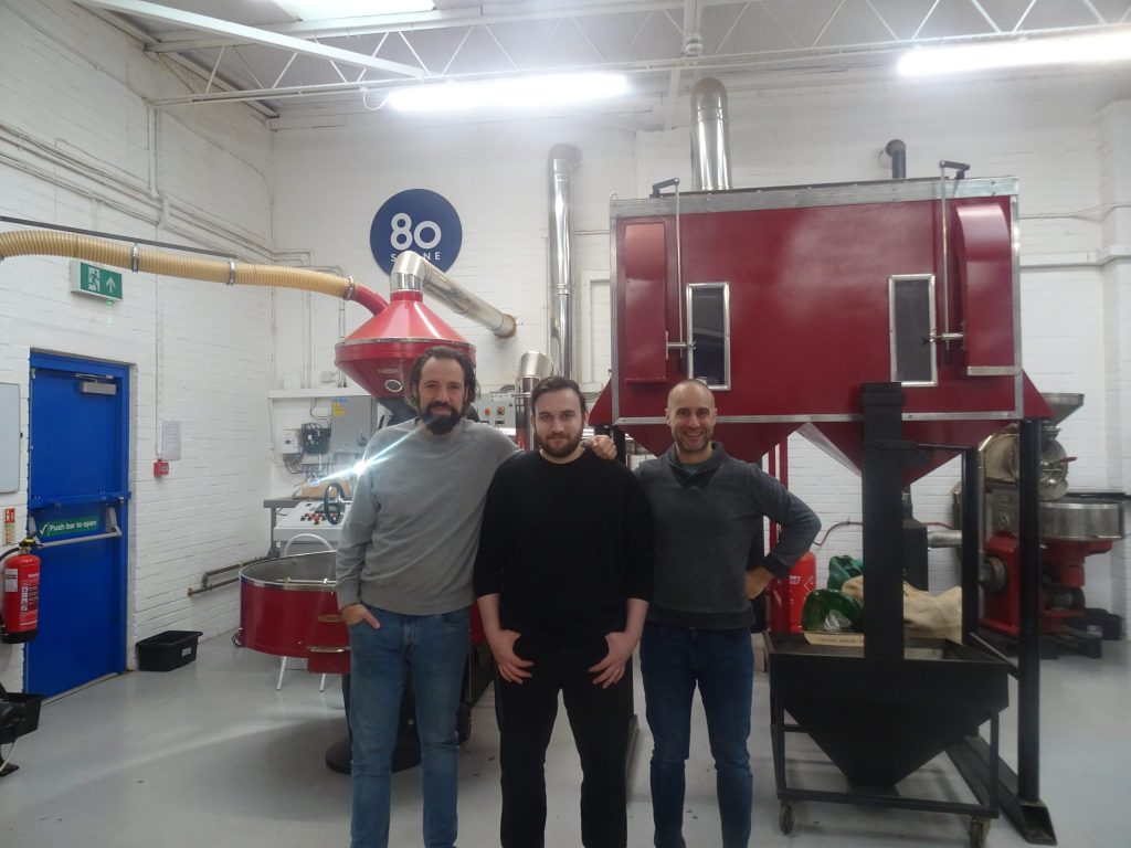 Three men pose in front of a red coffee roaster