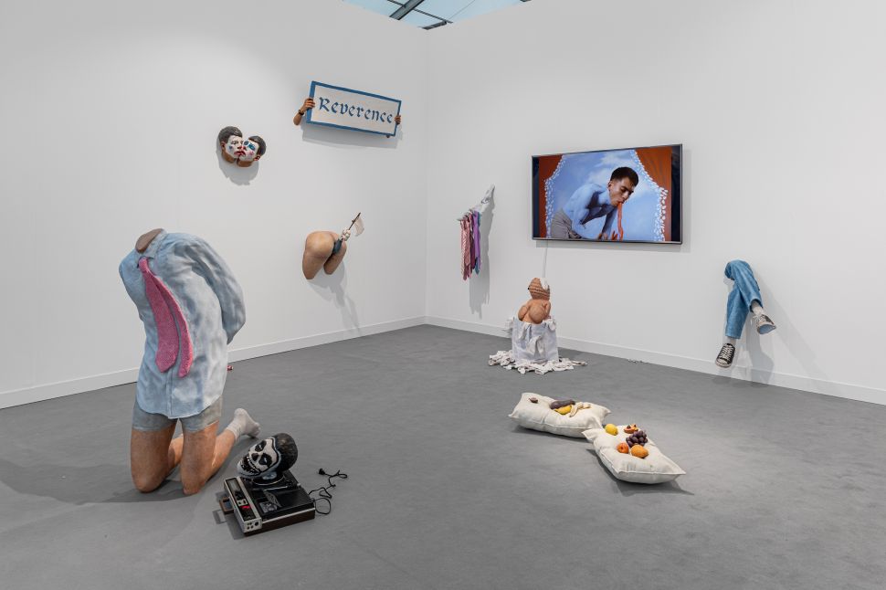 Installation view of LLANO’s booth at Frieze London 2025 featuring a surreal multimedia installation: fragmented sculptural body parts emerging from walls and floor, soft fabric sculptures resembling food on pillows, a kneeling headless figure beside an old tape recorder, and a video of a painted performer displayed on a wall monitor, all arranged in a sparse white-walled space.