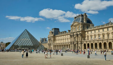 A triangular shaped glass building next to a larger, classical stone building.