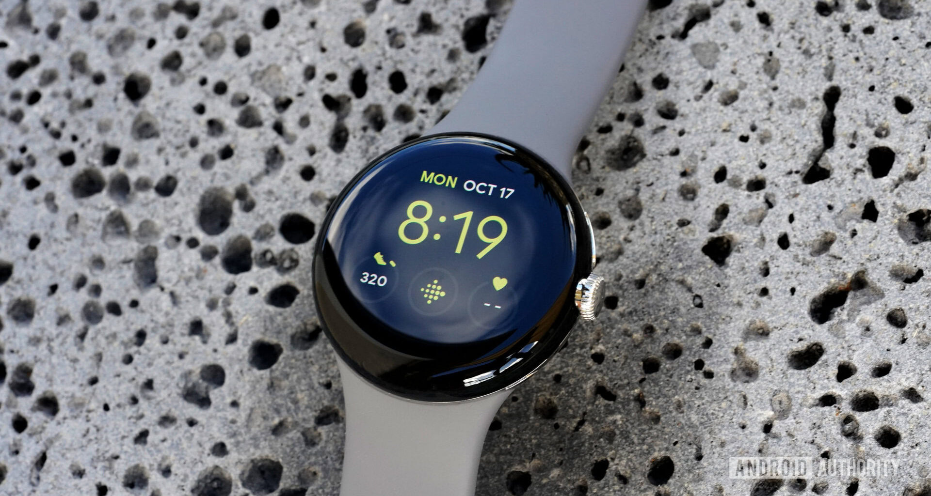 A Google Pixel Watch rests on a lava rock displaying the Utility watch face with a Fitbit shortcut.
