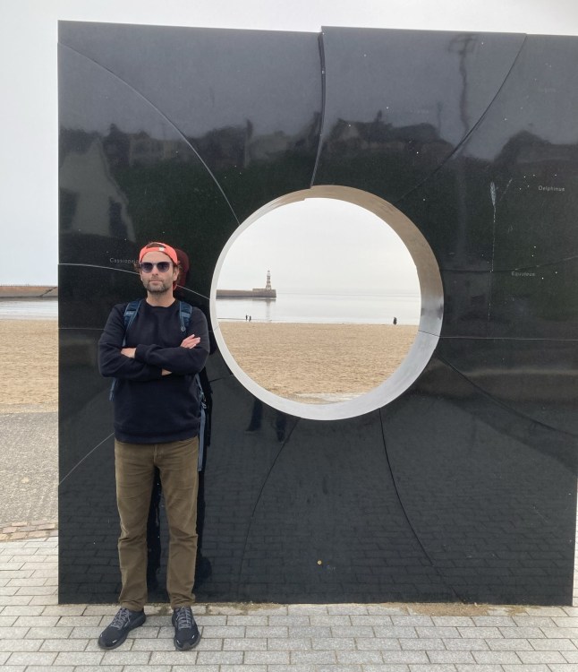 Ben Aitken in front of a sculptural wall on a beachfront in Sunderland