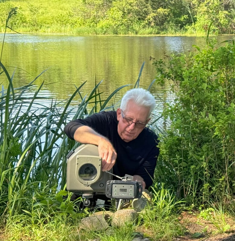 A person with white hair, wearing glasses and a black shirt, adjusts equipment near a lake surrounded by green plants and tall grass on a sunny day.