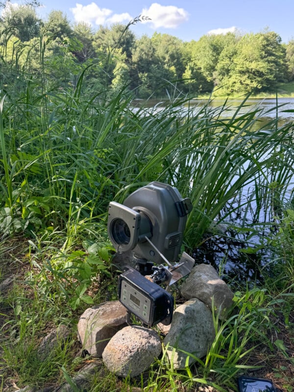 A wildlife camera with a protective case is mounted on rocks by a grassy riverbank. Tall green grass and leafy trees surround a calm body of water under a sunny, blue sky with scattered clouds.