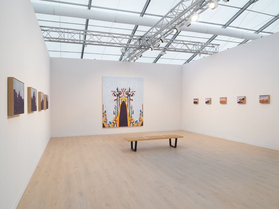 Installation view of Pace Gallery’s solo presentation of William Monk at Frieze London 2025, featuring a large central painting with surreal architectural forms on the back wall, flanked by smaller landscape-like canvases on both side walls, with a simple wooden bench placed in the center of the light-filled booth.