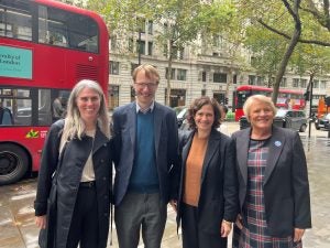 Four people stand together on a city street in front of a red double-decker bus.