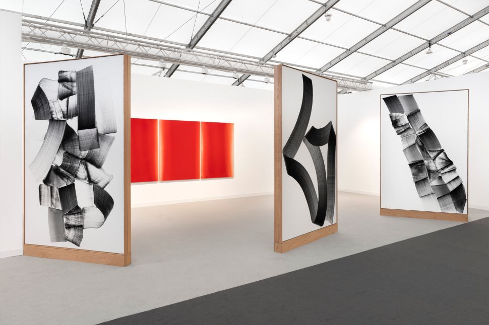 Fair view with three large freestanding panels featuring bold black abstract brushstroke compositions, framing a bright red triptych on the back wall in a minimal white booth.