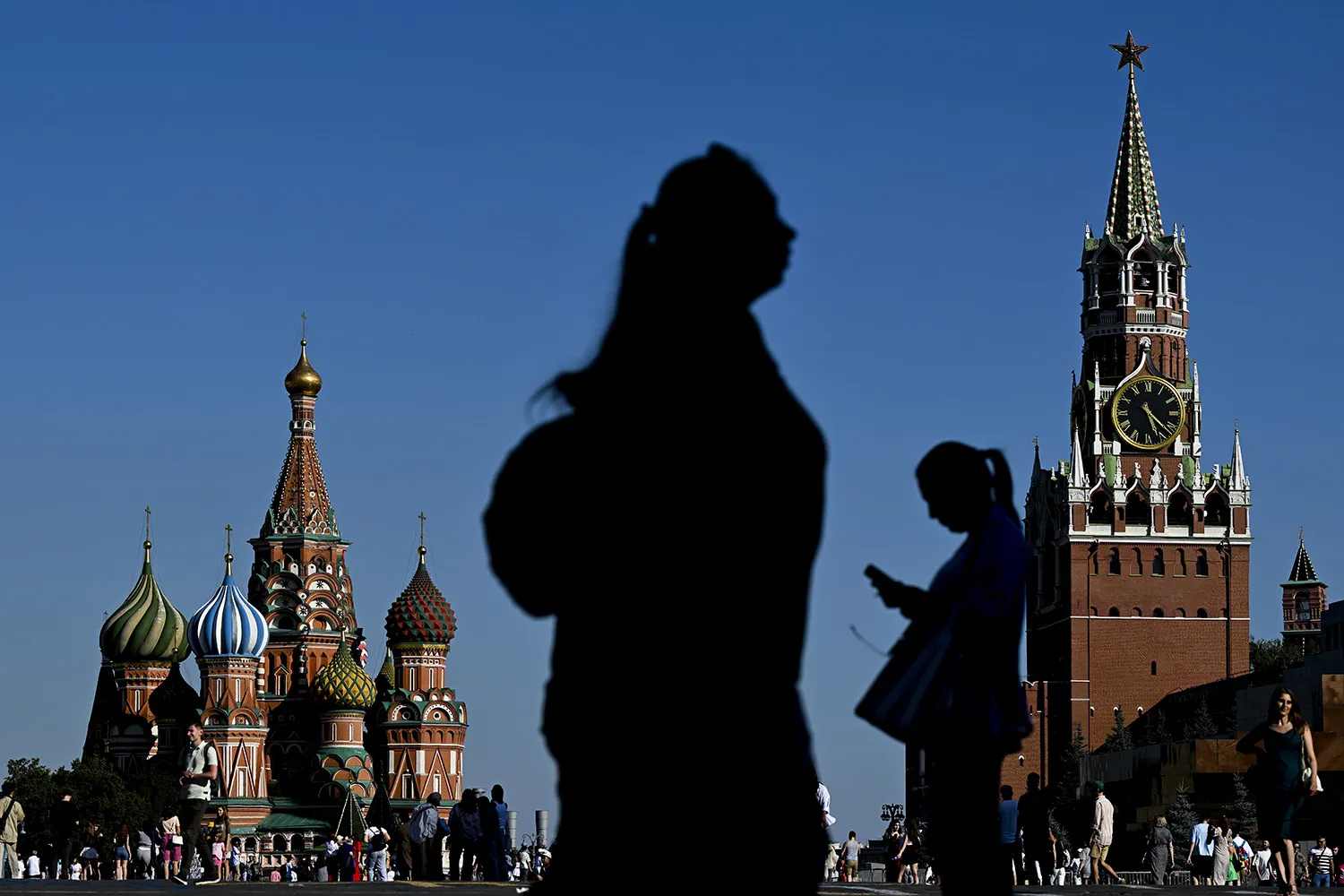 In the foreground is the silhouette of a woman walking in one direction, with the silhouette of another woman, facing the opposite direction and looking at her phone, in the midground. In the background are the towers of the St. Basil Cathedral and the Spasskaya clock tower in the historic Red Square in Moscow, and crowds of people milling through the square.