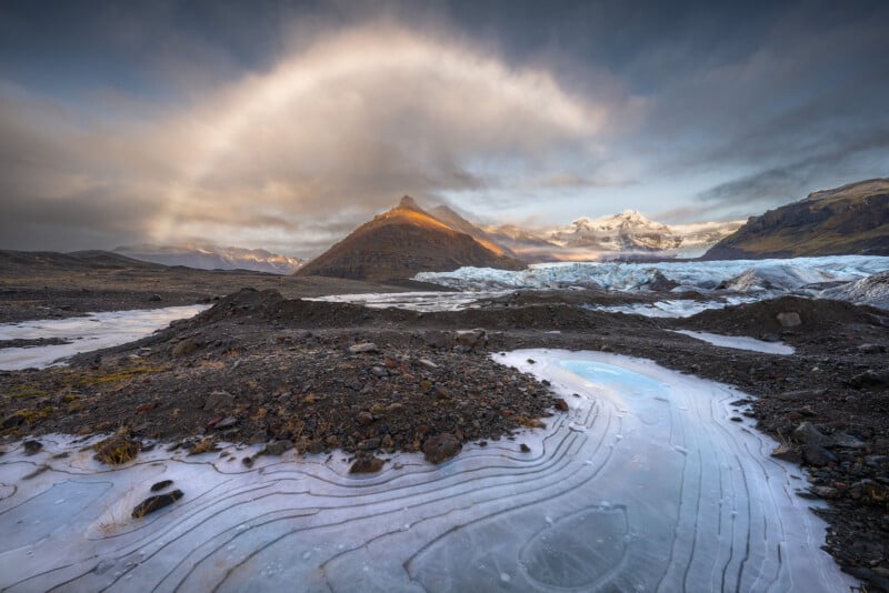 A frozen landscape features icy ground with curved lines, rocky terrain, a glacier, and a distant mountain bathed in sunlight under a cloudy sky with a faint rainbow-like arc.