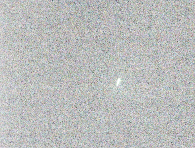 A grainy, noisy image with a mostly gray background and a single short white streak near the center, resembling a small bright light or object amidst heavy visual static.