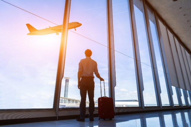 A man with a suitcase watching a plane fly past a large airport window.