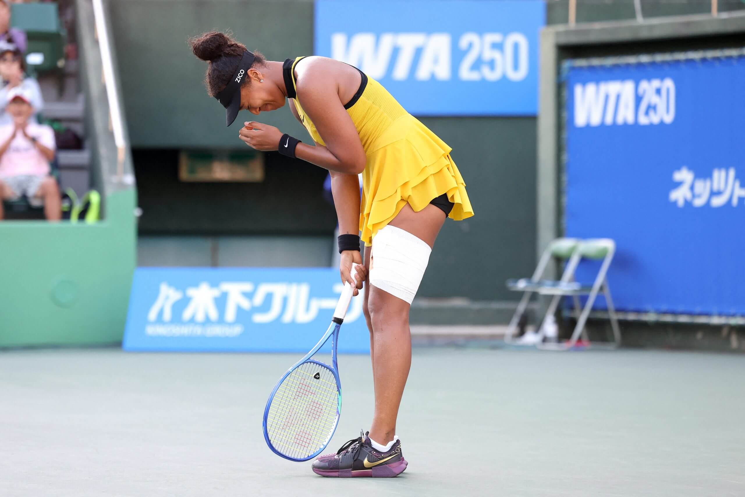 Naomi Osaka bends over in a mixture of pain and triumph while holding her tennis racket. She wears a yellow dress with a large white bandage on her upper left leg.