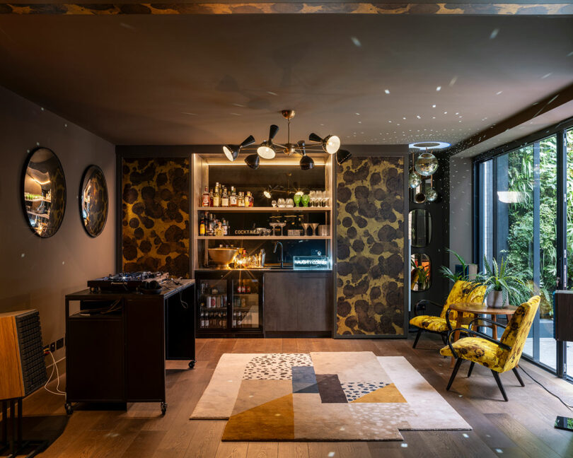 Modern home bar with patterned walls, open shelving, cocktail supplies, yellow chairs, a geometric rug, and large glass doors overlooking greenery.