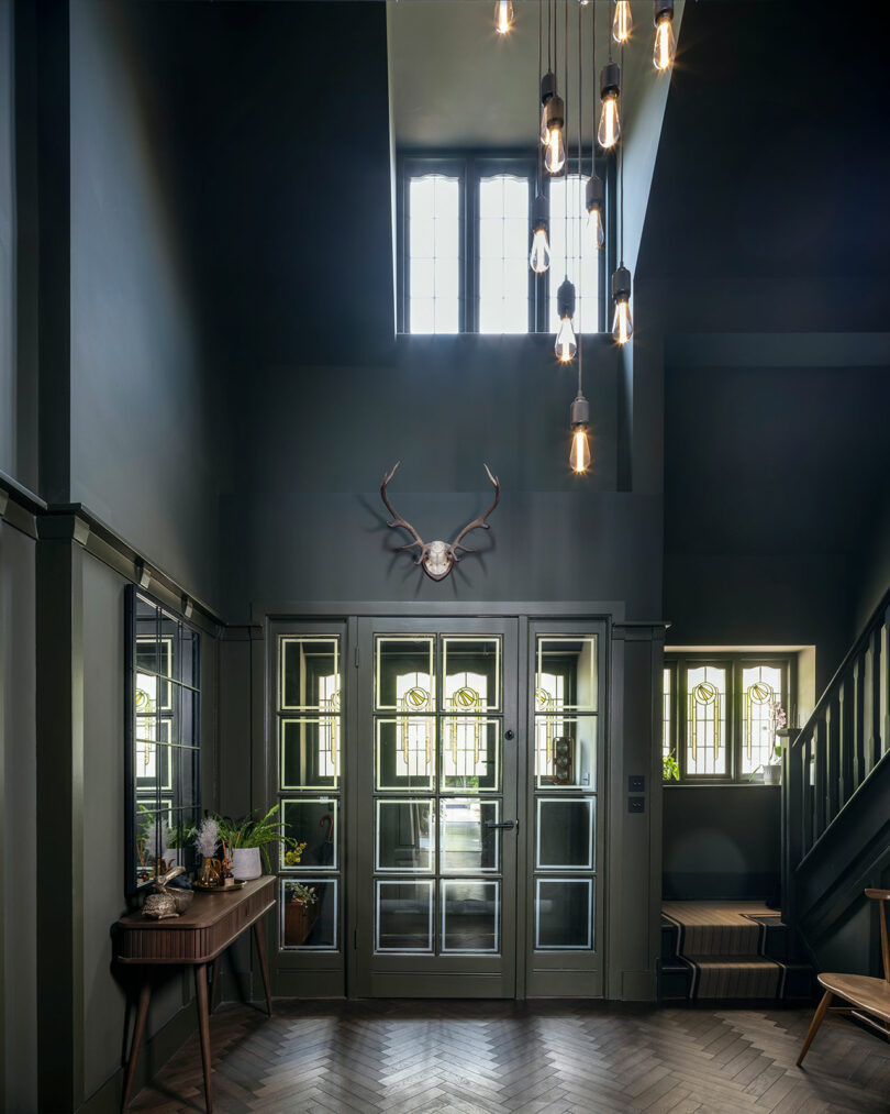 A modern hallway with dark walls, glass double doors, a mounted deer skull with antlers, pendant lights, a side table with plants, and a staircase.
