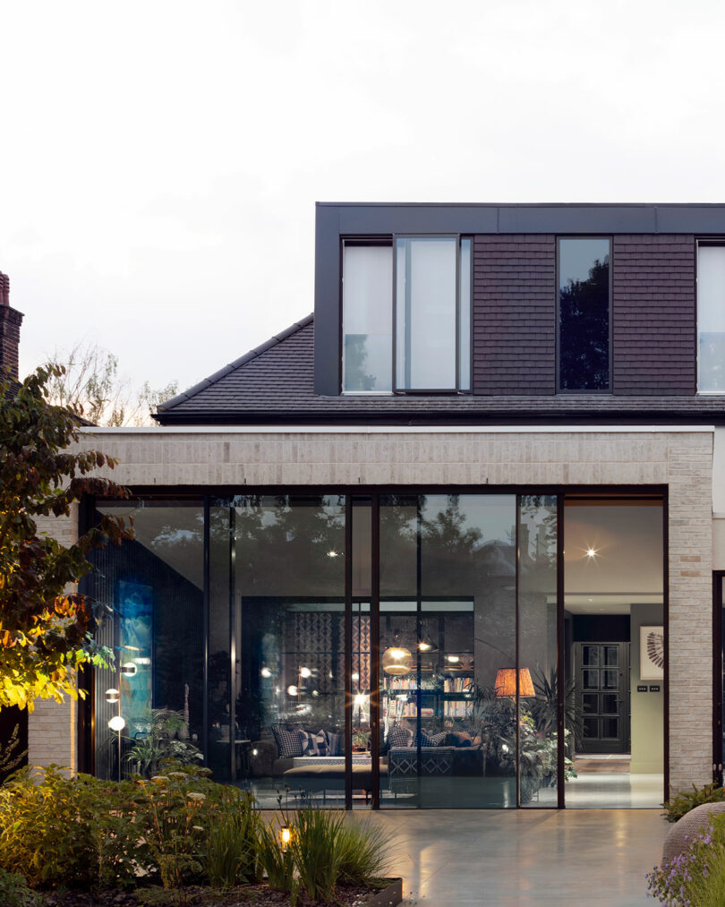 Modern two-story house with large floor-to-ceiling glass windows and visible interior furnishings, surrounded by landscaped greenery.