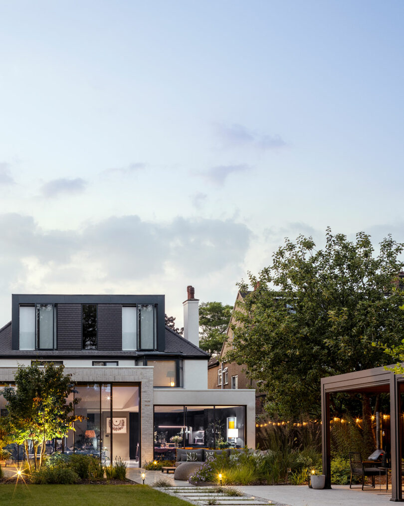 Modern two-story house with large windows, a well-kept garden, and outdoor seating area at dusk; sky is clear with a few clouds.