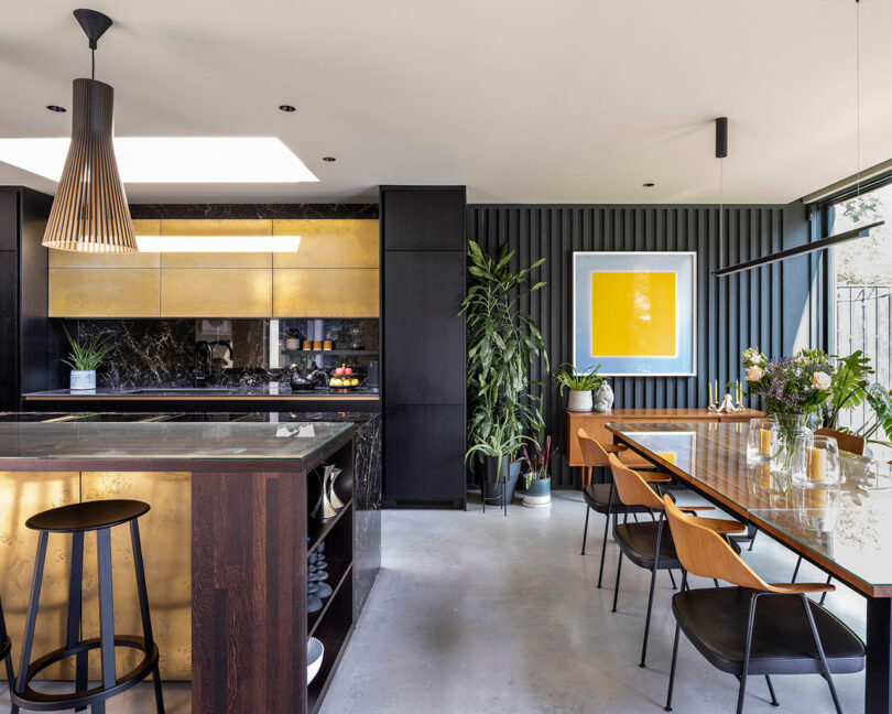 Modern kitchen and dining area featuring dark cabinetry, gold accents, a marble countertop, indoor plants, and a large yellow framed artwork on the wall.
