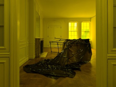 A room lit yellow with a sculpture on its floor. The sculpture resembles a tipped-over armature with a latex drape over it.