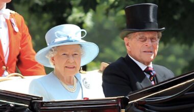 An elderly woman in a light blue outfit and hat sits beside an elderly man in a black suit and top hat in an open carriage, with another person in a red coat partially visible behind them. Trees are in the blurred background.