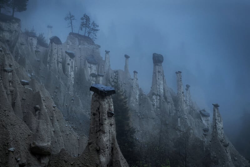 Tall, slender rock formations topped with large stones rise from a misty, forested hillside, shrouded in blue-grey fog. Sparse trees grow atop the cliffs and along the slopes, creating an eerie, mysterious landscape.
