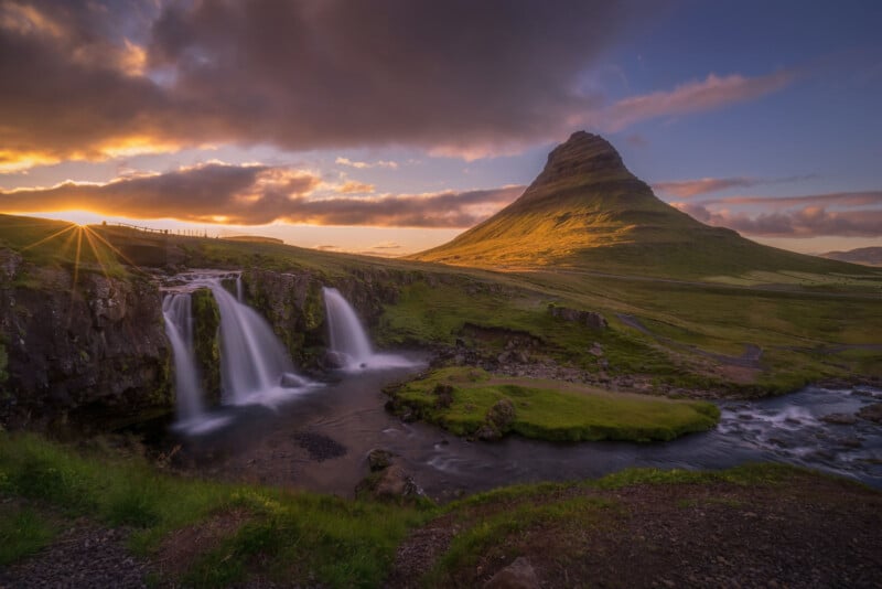 A scenic landscape at sunset with a mountain in the background, a river and small waterfalls flowing over green mossy rocks, and sunlight streaming through dramatic clouds.