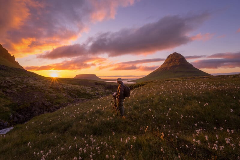 A person with a backpack and tripod stands in a field of wildflowers at sunset, facing a distinctively shaped mountain under a colorful sky with dramatic clouds.