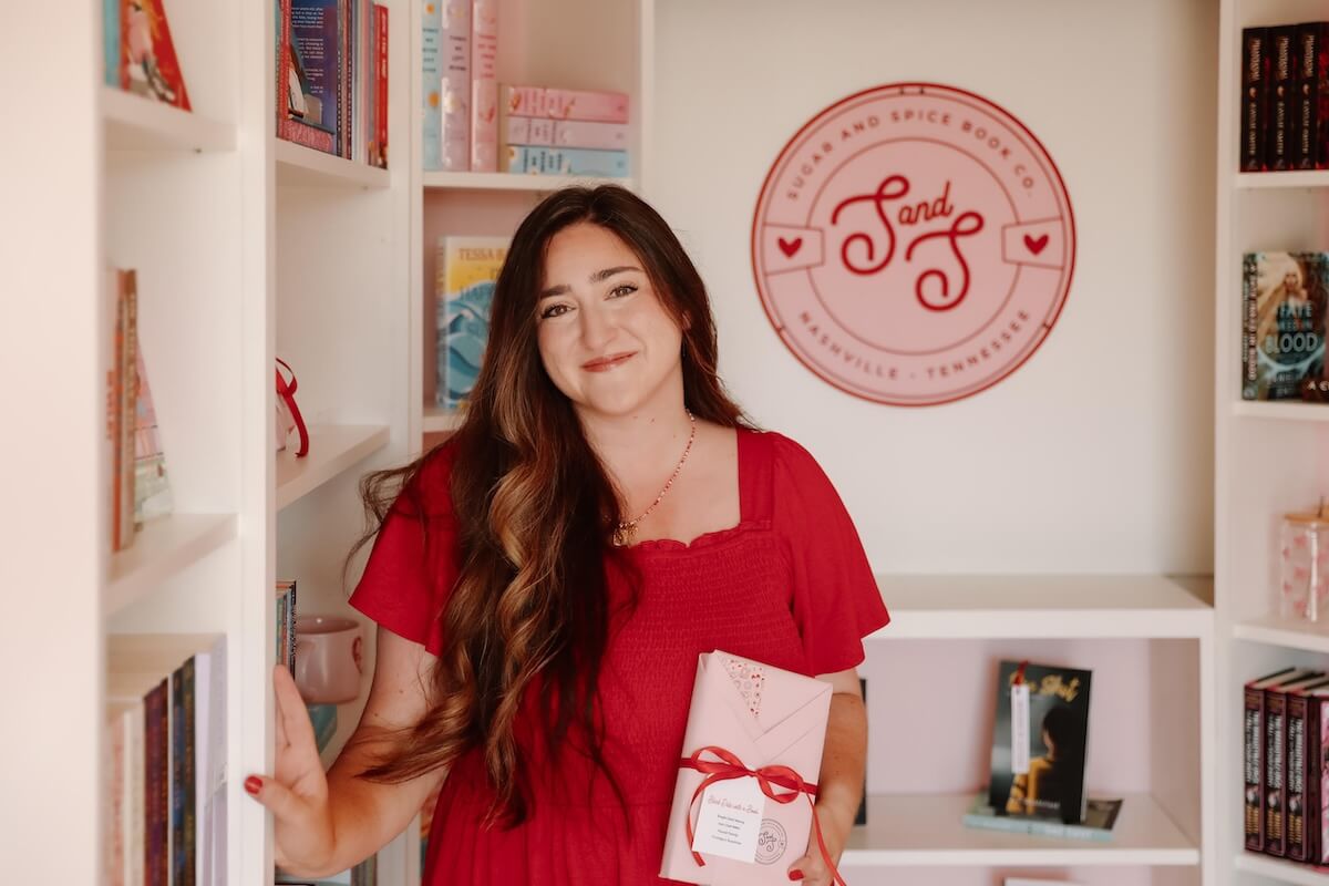 She's Behind Nashville's Sugar and Spice Book Co. - 3 Christina Duran, in a red dress, stands in a bookstore holding a wrapped book. Shelves of books and a wall sign reading “Sugar And Spice Book Co” are visible in the background.