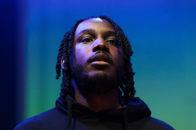 Shocka is looking up in a headshot. The background is blue and green. He is wearing a dark hoody and has a serious expression