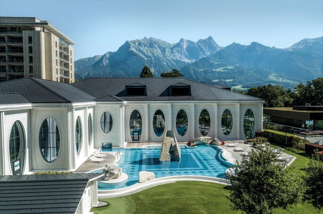 A high view on an outdoor pool with a sculpture in the middle in front of a white building at Grand Resort Bad Ragaz. There are mountains behind in the distance under a blue sky.