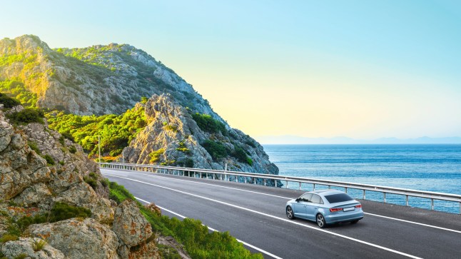 A car driving down a scenic Spanish road, surrounded by blue sea and rocky cliffs.