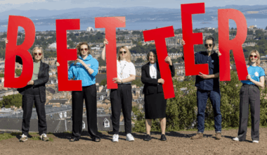 Six people standing in a horizontal row on a hilltop, each person holding a letter sign to spell out the word 'Better'.