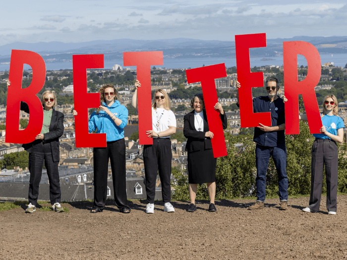 Six people standing in a horizontal row on a hilltop, each person holding a letter sign to spell out the word 'Better'.