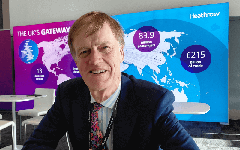 Stephen Timms smiling, in a suit and tie, sitting, with a map of the world behind him