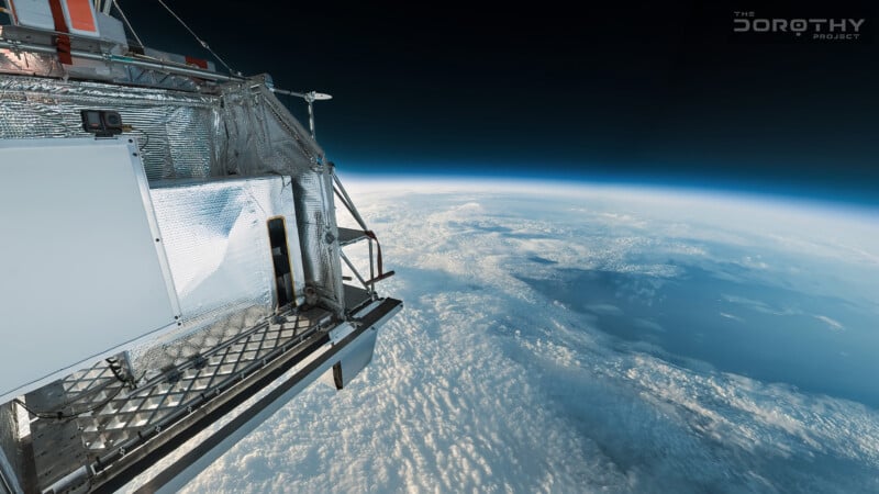 View from a metallic space probe or high-altitude balloon above Earth, showing part of the craft and a vast layer of clouds, with Earth's curvature and the dark expanse of space in the background.