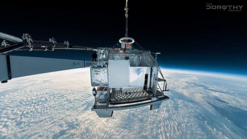 A scientific payload covered in reflective foil is suspended above Earth, attached to a cable, with clouds and the curve of the planet visible below against the blackness of space.