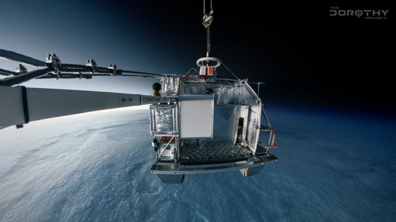 A metallic scientific instrument floats above Earth, suspended by cables with a vast layer of clouds and the planet's horizon below, capturing data as part of the Dorothy Project.