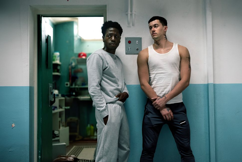 Two men stand in a prison or institutional hallway, one wearing gray sweats and the other a white tank top, looking at each other with tense expressions.