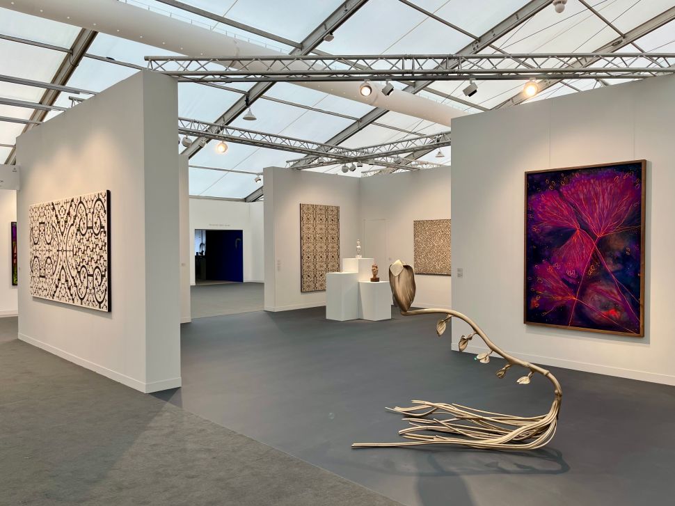 Booth with large abstract patterned paintings and a metallic plant-like floor sculpture extending across the space, under bright exhibition lighting.