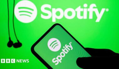 The silhouette of a hand holds a phone with the green and white Spotify logo on it. In the background hang some black wired earbuds.