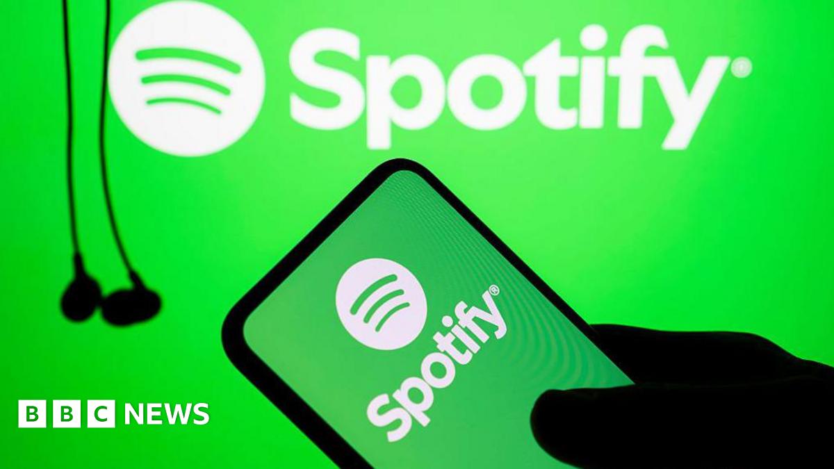 The silhouette of a hand holds a phone with the green and white Spotify logo on it. In the background hang some black wired earbuds.