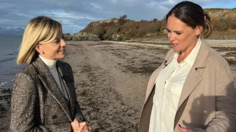 Susie Wolff, a woman with medium-length blonde hair, is standing on a beach somewhere rural. The sea and some distant land can be seen in the background. There is sand just behind her. She is wearing a black and white tweed checked jacket, white-polo-neck, and pearl ear-rings. She is smiling and it is sunny. She is talking to Laura Miller, who has short, dark, hair and is wearing a beige coat and cream blouse.
