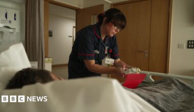 Image of a care staff member at a hospice bed which has a patient in it. She is wearing blue overalls and is looking at medication.