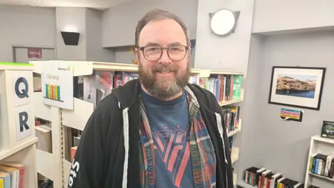 Stuart Neville is wearing glasses and has brown hair and a beard. 
He is wearing a black hoodie and a multi-coloured shirt and blue top. 
He is standing in the middle a library.