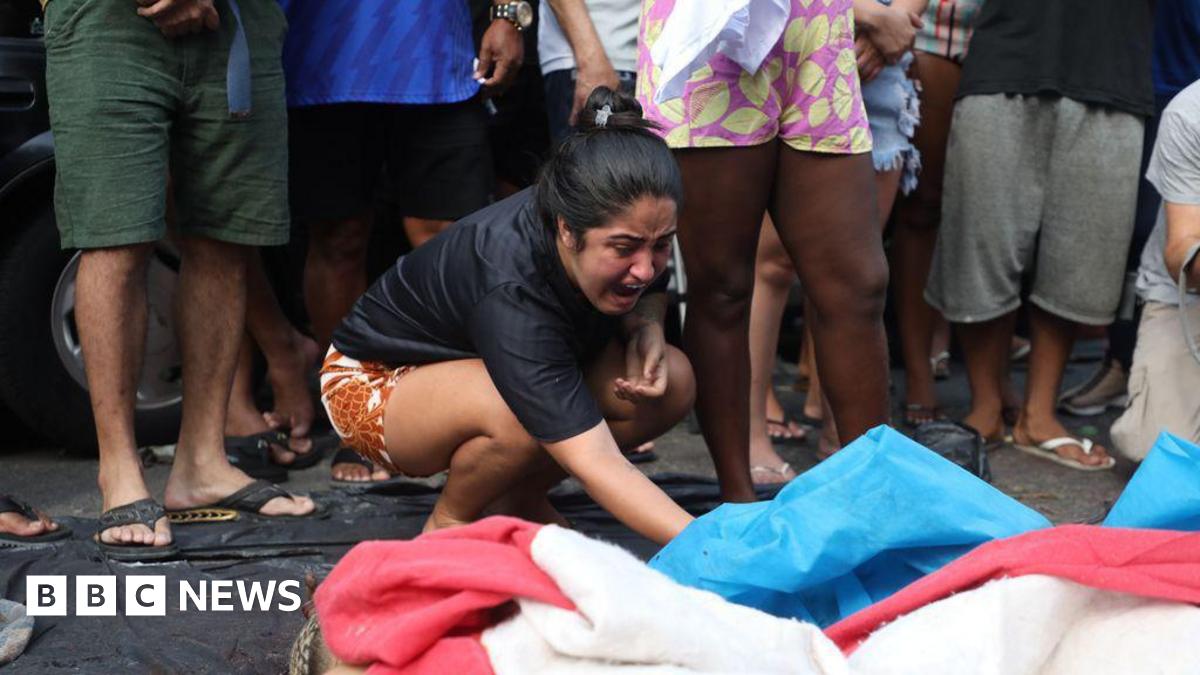 A woman lifts a blue tarpaulin which covers a body. She appears to be crying out. The legs of bystanders can be seen behind her.