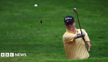 A golfer with a yellow tshirt and black cap striking the ball in the air.