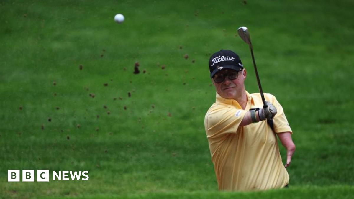 A golfer with a yellow tshirt and black cap striking the ball in the air.